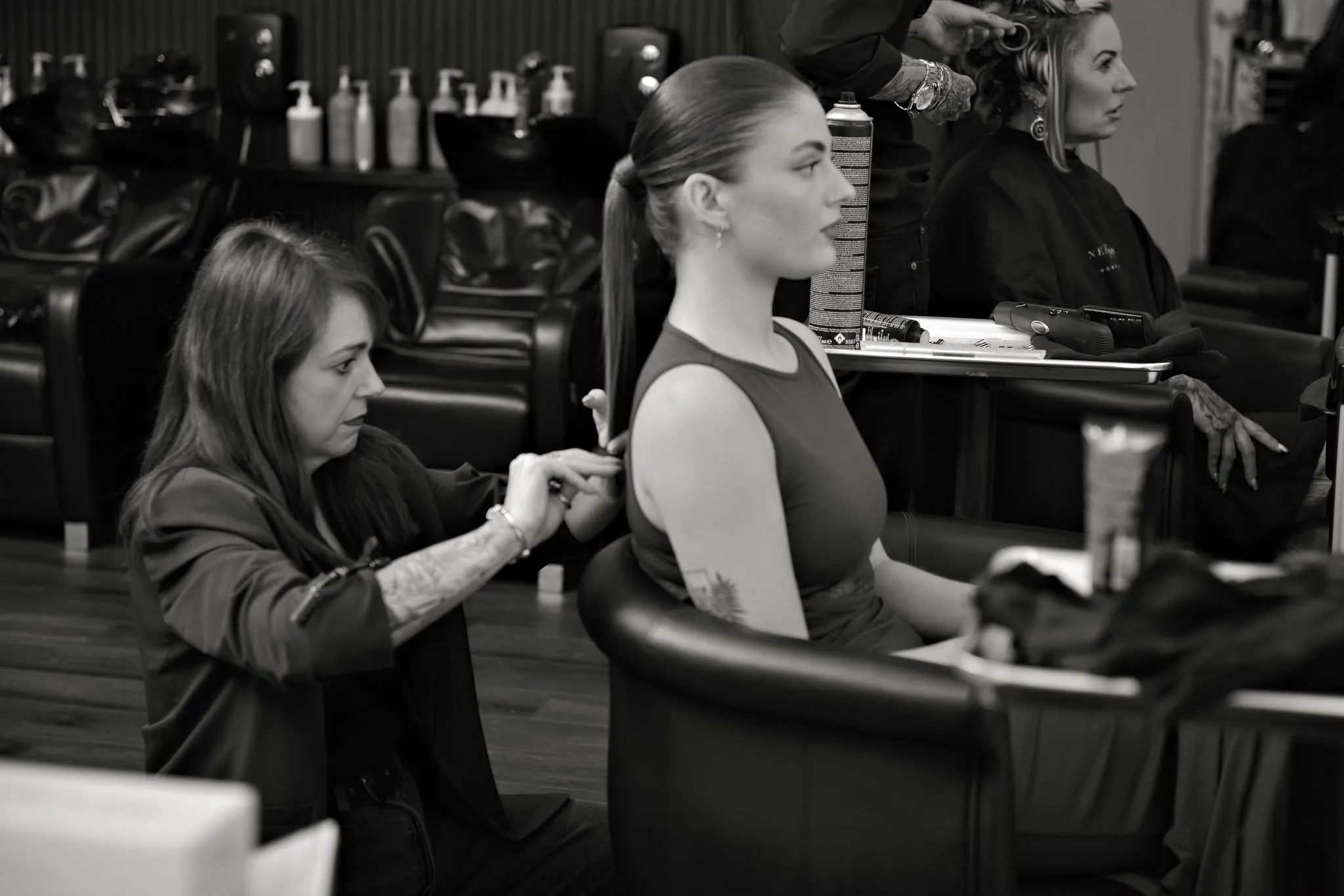 Katie working on a client's sleek high ponytail, with a second client in pin curls and the black leather wash basins behind