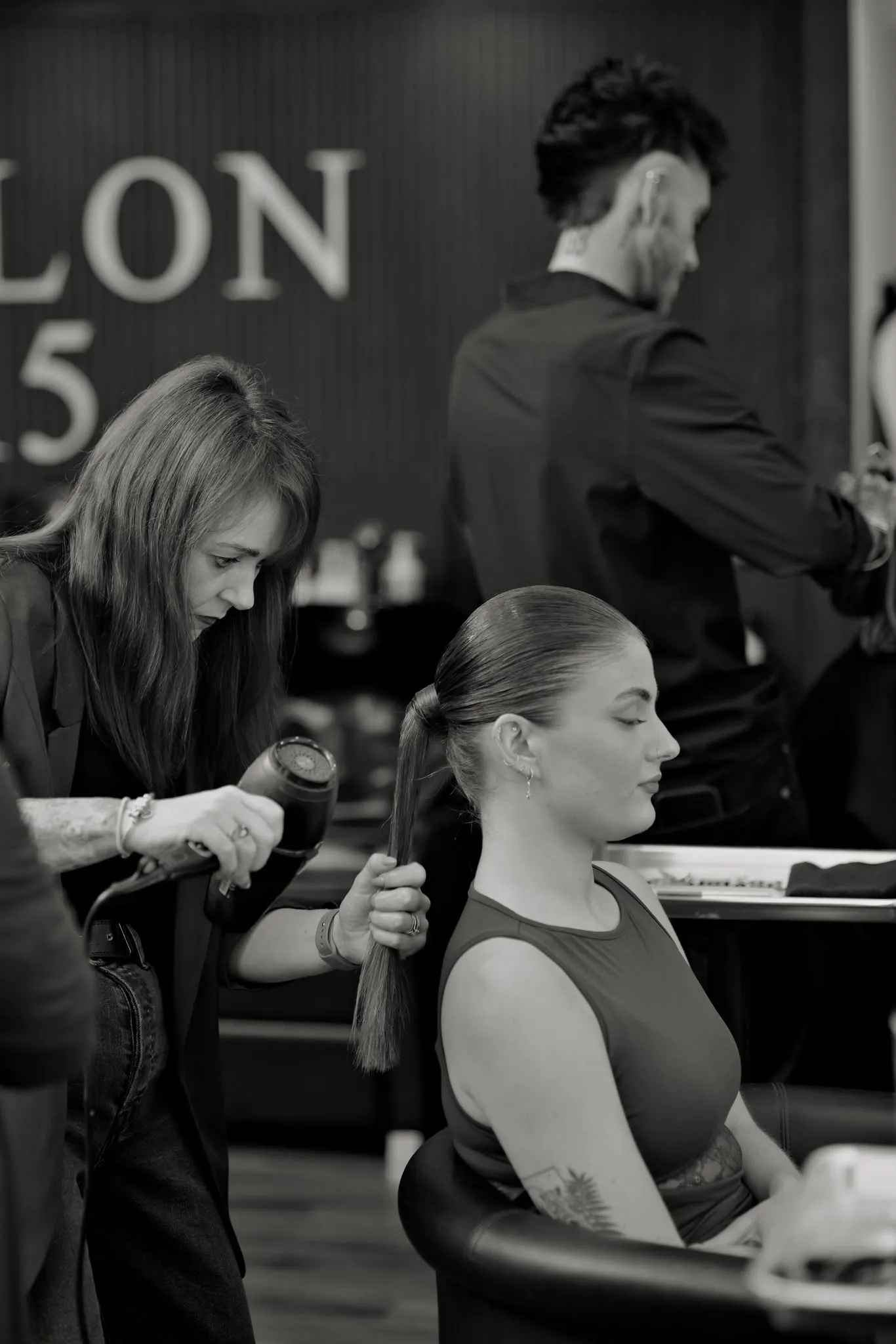 Katie blow-drying a client's sleek ponytail in front of the SALON 15 wall, with AJ working behind